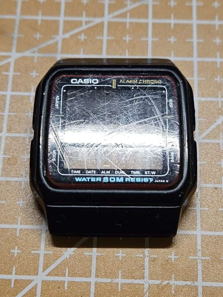 Casio AE-11W black resin watch case at slight angle showing scratched Alarm Chrono crystal before restoration