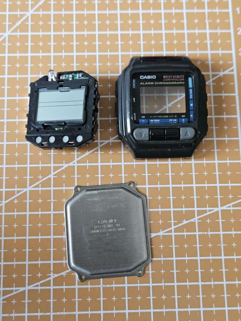 Casio CMD-30 fully disassembled showing inner module with LCD, outer case, and stainless steel case back on cutting mat
