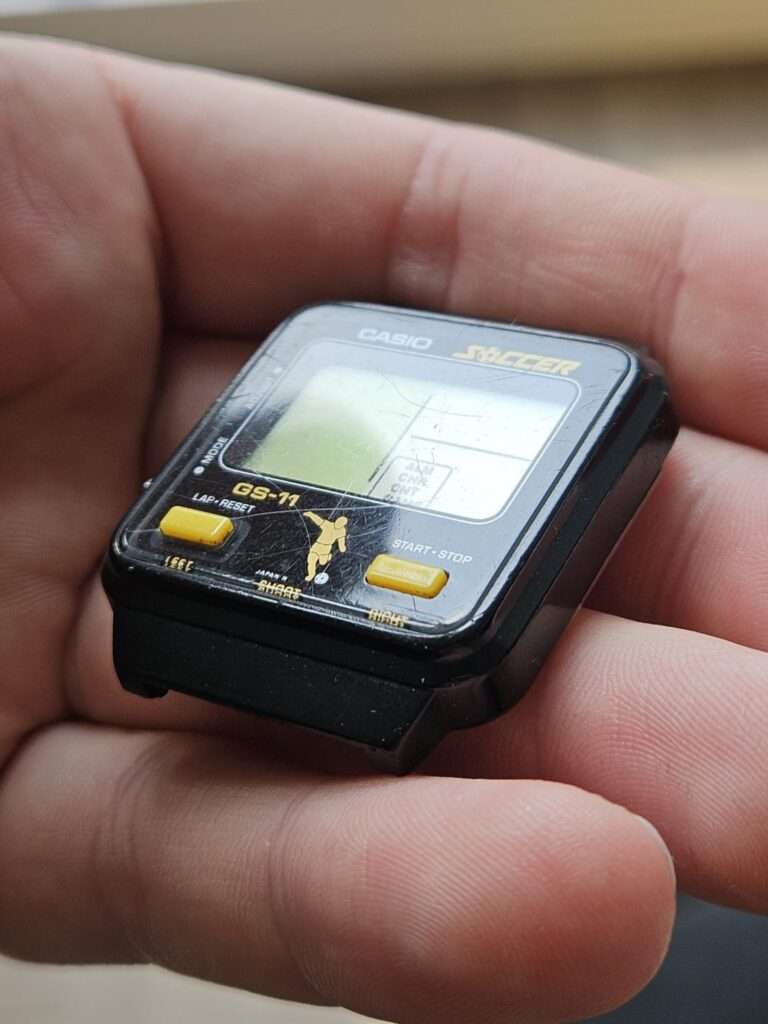 Casio GS-11 Soccer game watch held in hand showing front face with LCD and yellow buttons before restoration