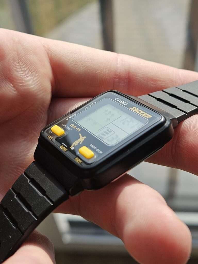 Side-oriented flat-lay of the post-restoration Casio GS-11 Soccer game watch on an orange grid mat, highlighting the refurbished black bracelet links, clean case profile, and a fully legible LCD displaying an ongoing soccer game with time and score readouts.