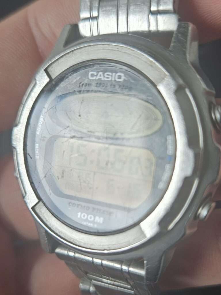Casio CGW-500 Cosmo Phase digital watch held in hand, close-up of heavily scratched mineral crystal before restoration