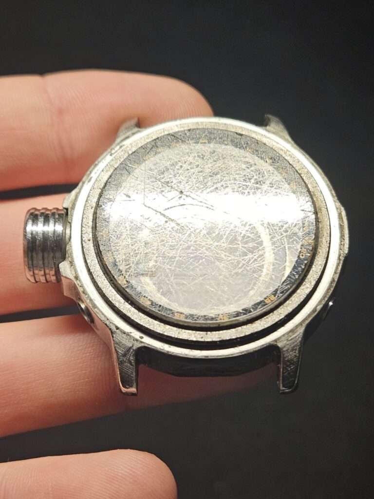 Casio watch case held in fingers showing a heavily scratched mineral crystal and crown